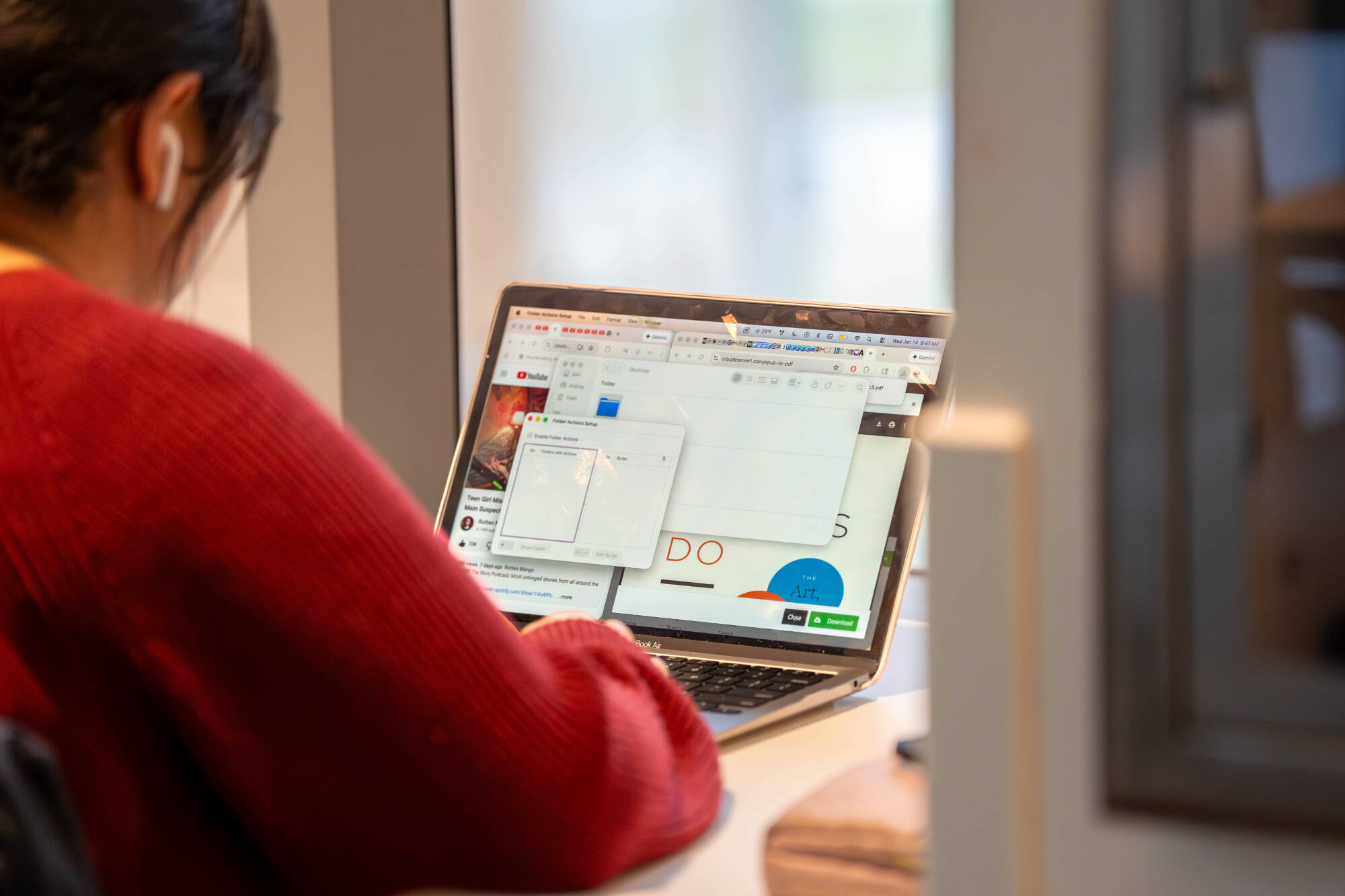 A person in a red sweater works on a laptop with earbuds in a modern work environment. The screen displays a YouTube page and colorful graphic elements.